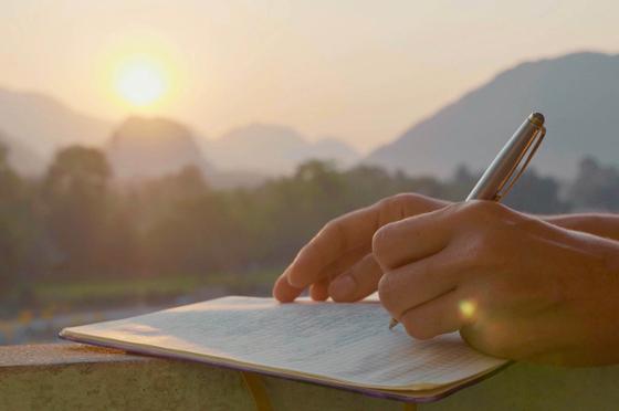 Photo of a person's hands, as they write in a notebook with a sunset in the background