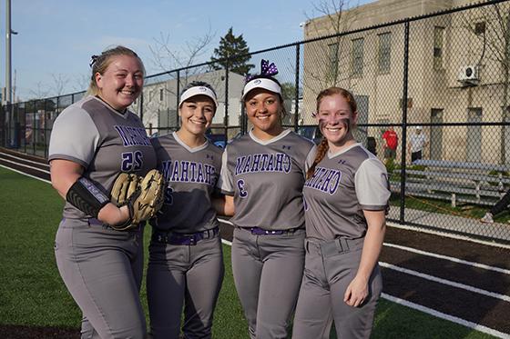 Photo of four Chatham University female softball players pose together in their gray and purple uniforms before a game