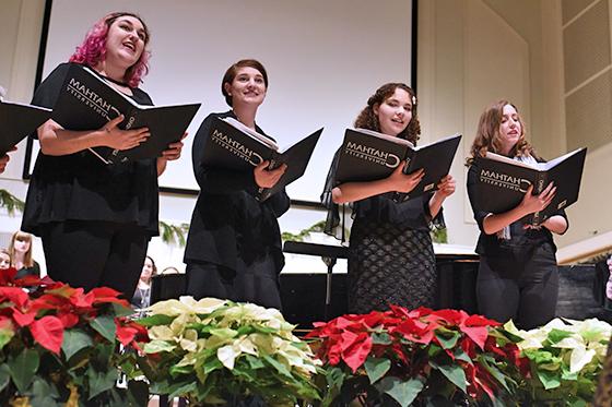 Photo of four Chatham University students singing on stage in a choir while holding music binders and standing in front of an orchestra